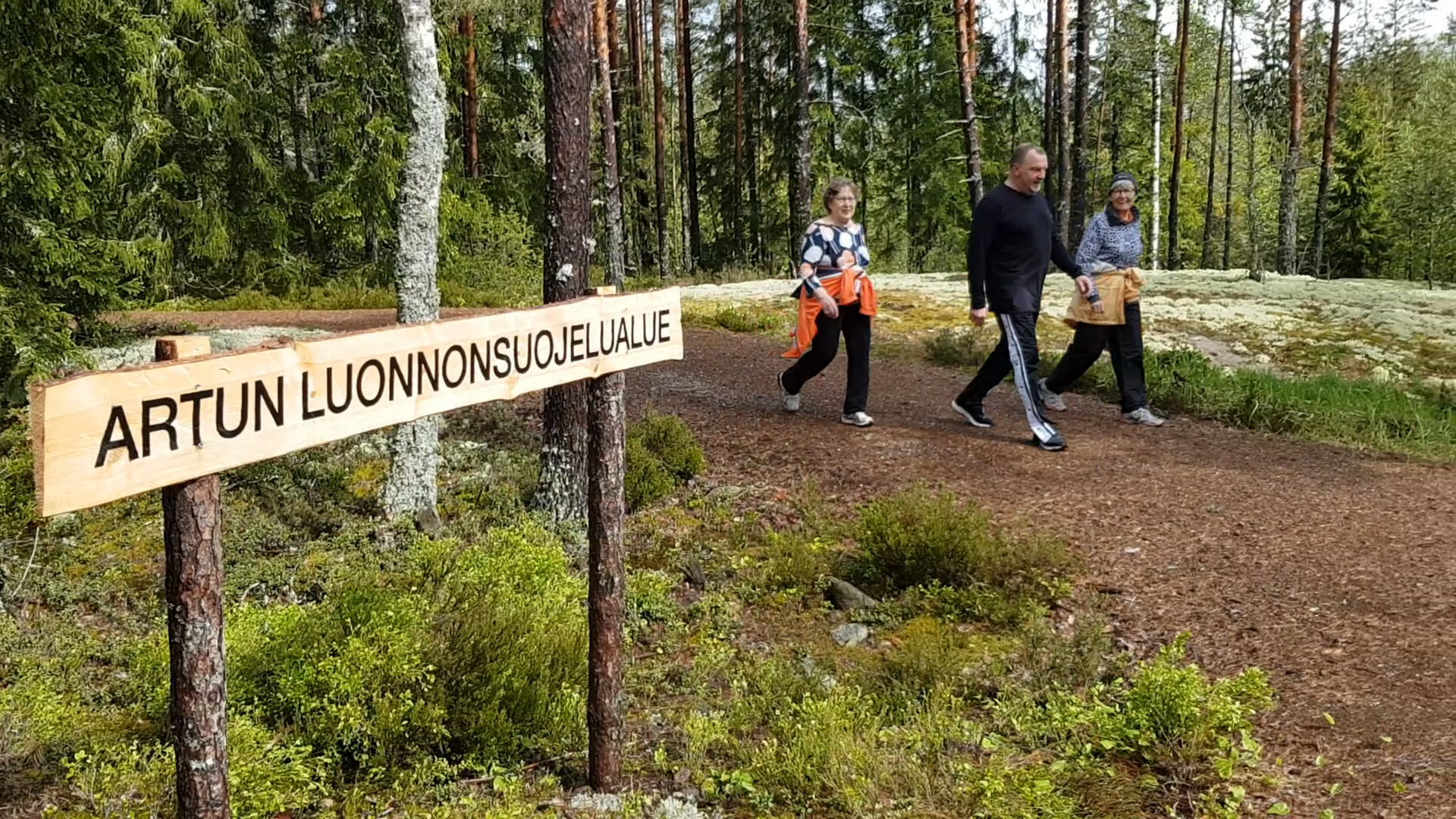 Puinen Artun luonnonsuojelualueen kyltti, jonka ohi kolme kävelijää on kävelemässä.