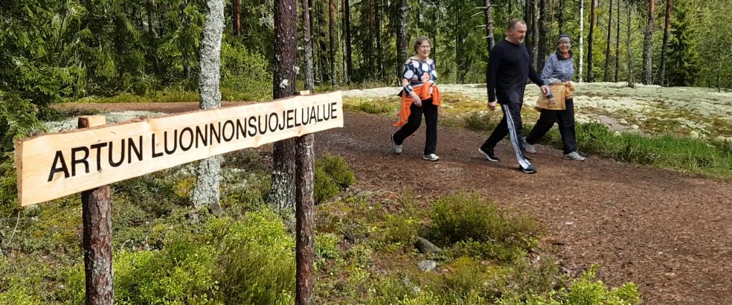 Puinen Artun luonnonsuojelualueen kyltti, jonka ohi kolme kävelijää on kävelemässä.